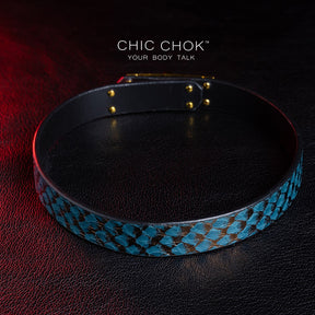 chicchok [Sapphire Moonlight] choker featuring a teal snakeskin-textured band with a gold-plated buckle clasp, displayed on a dark leather surface with dramatic red lighting accents, showcasing its luxurious and edgy design.