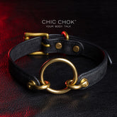 chicchok [Sinister] collar black leather choker featuring a central gold ring, gold-plated hardware, and adjustable buckle design, photographed on a textured black surface with warm lighting highlights